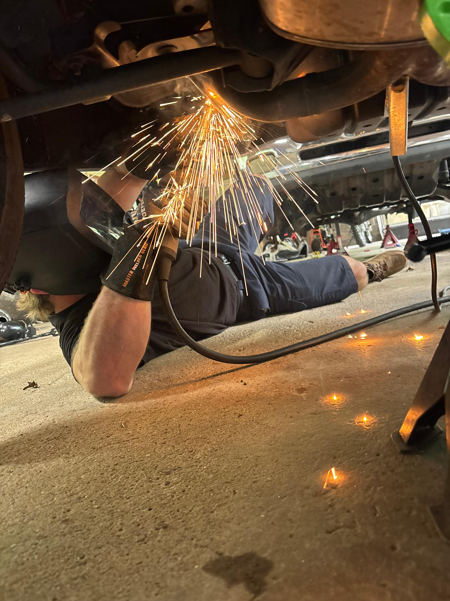 G23 Solutions technician MIG welding an exhaust repair from underneath a vehicle with sparks flying