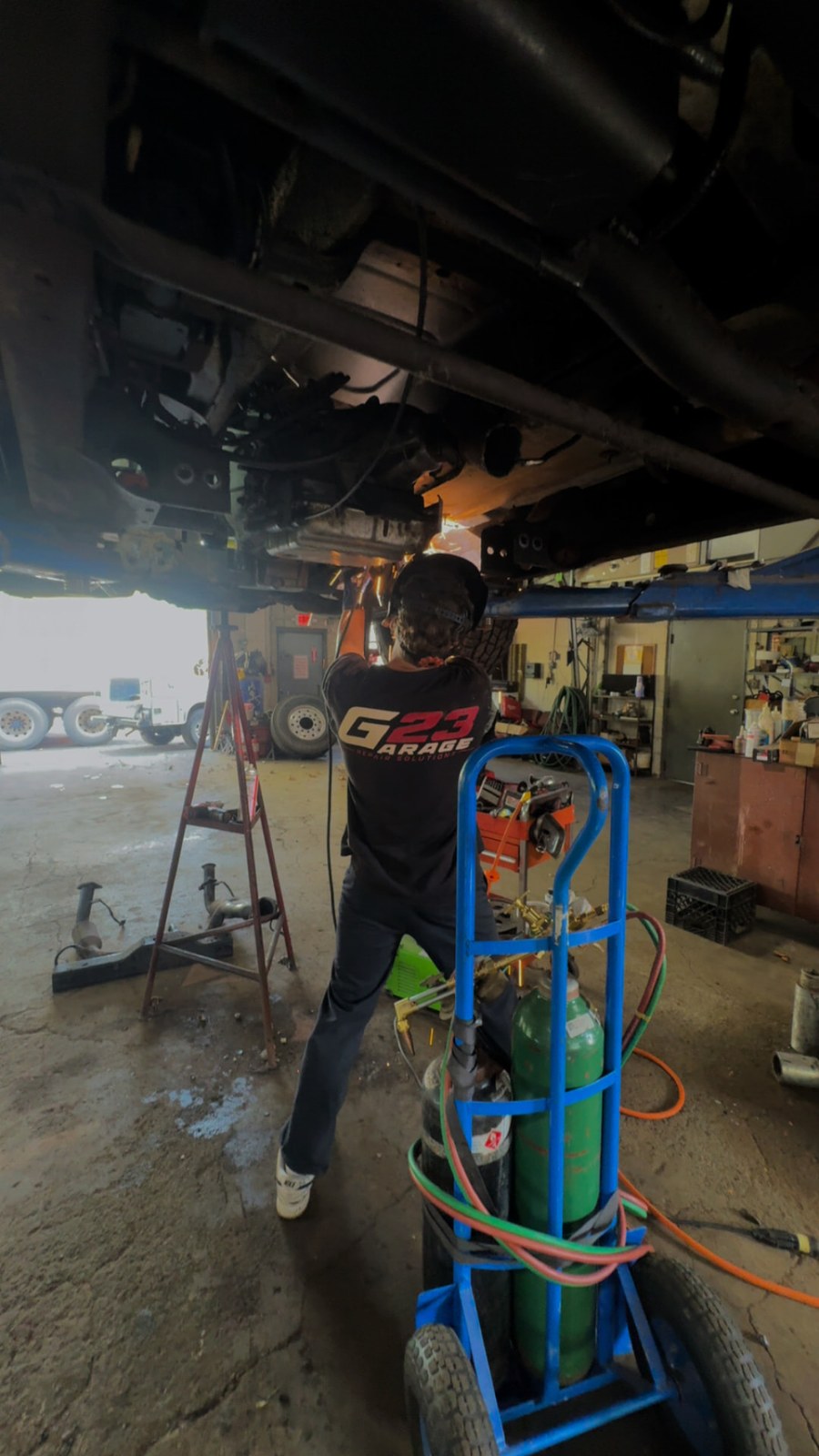 G23 technician in a G23 shirt cutting and welding under a raised vehicle in the shop, with a torch cart alongside