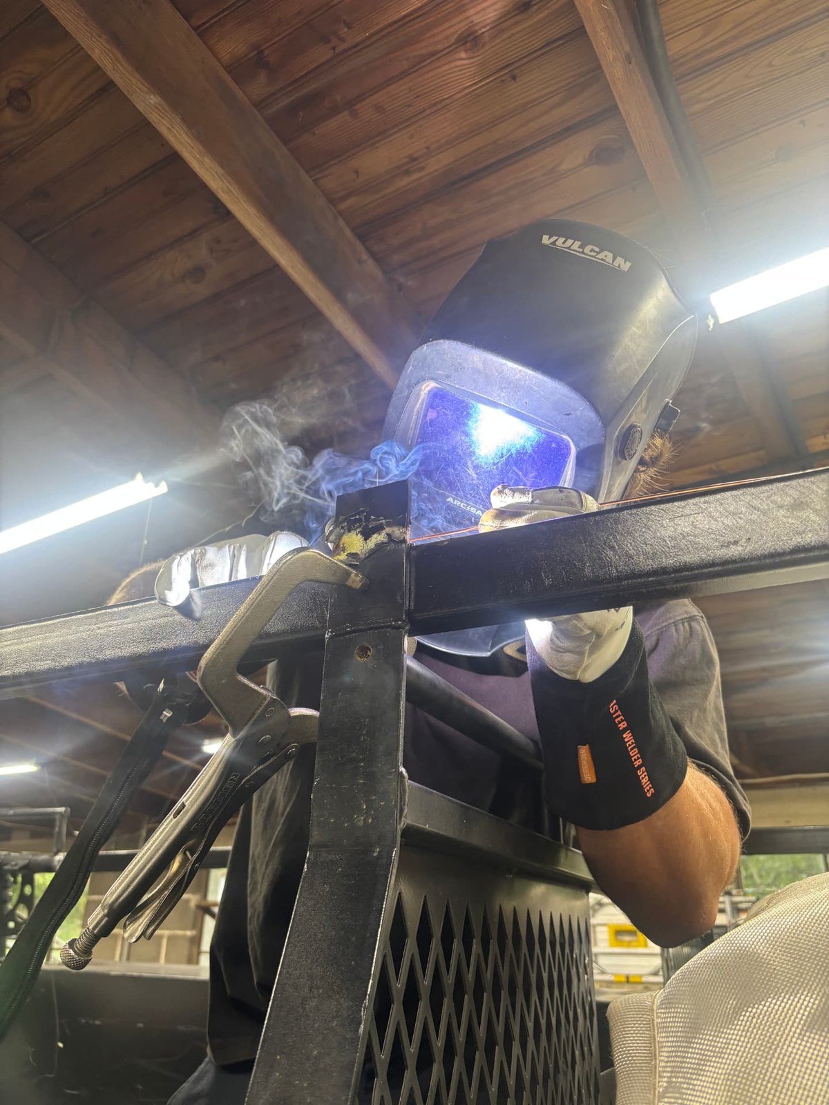 Welder in a welding helmet striking an arc on a steel trailer frame clamped in the shop